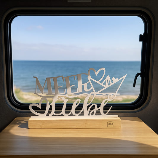 Weißer Deko-Aufsteller mit „Meer Liebe“-Schriftzug und Herz auf Holzsockel steht vor einem Fenster mit Blick auf das Meer im Camper oder Wohnmobil.
