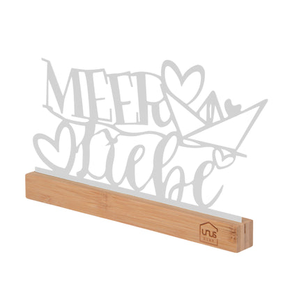 Weißer Aufsteller mit Schriftzug „Meer Liebe“ und Herz-Boot-Motiv aus Metall, schräg von vorn fotografiert, auf Holzsockel mit UNUS® HOME Logo.