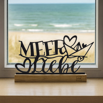 Schwarzer Aufsteller mit dem Schriftzug „Meer Liebe“ und Herz-Boot-Motiv auf Holzsockel steht auf einer Fensterbank mit Blick auf Strand und Meer.