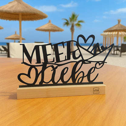 Schwarzer Deko-Aufsteller mit Schriftzug „Meer Liebe“ auf Holzsockel steht auf einem Tisch vor Strandkulisse mit Palmen, Sonnenschirmen und Meer.
