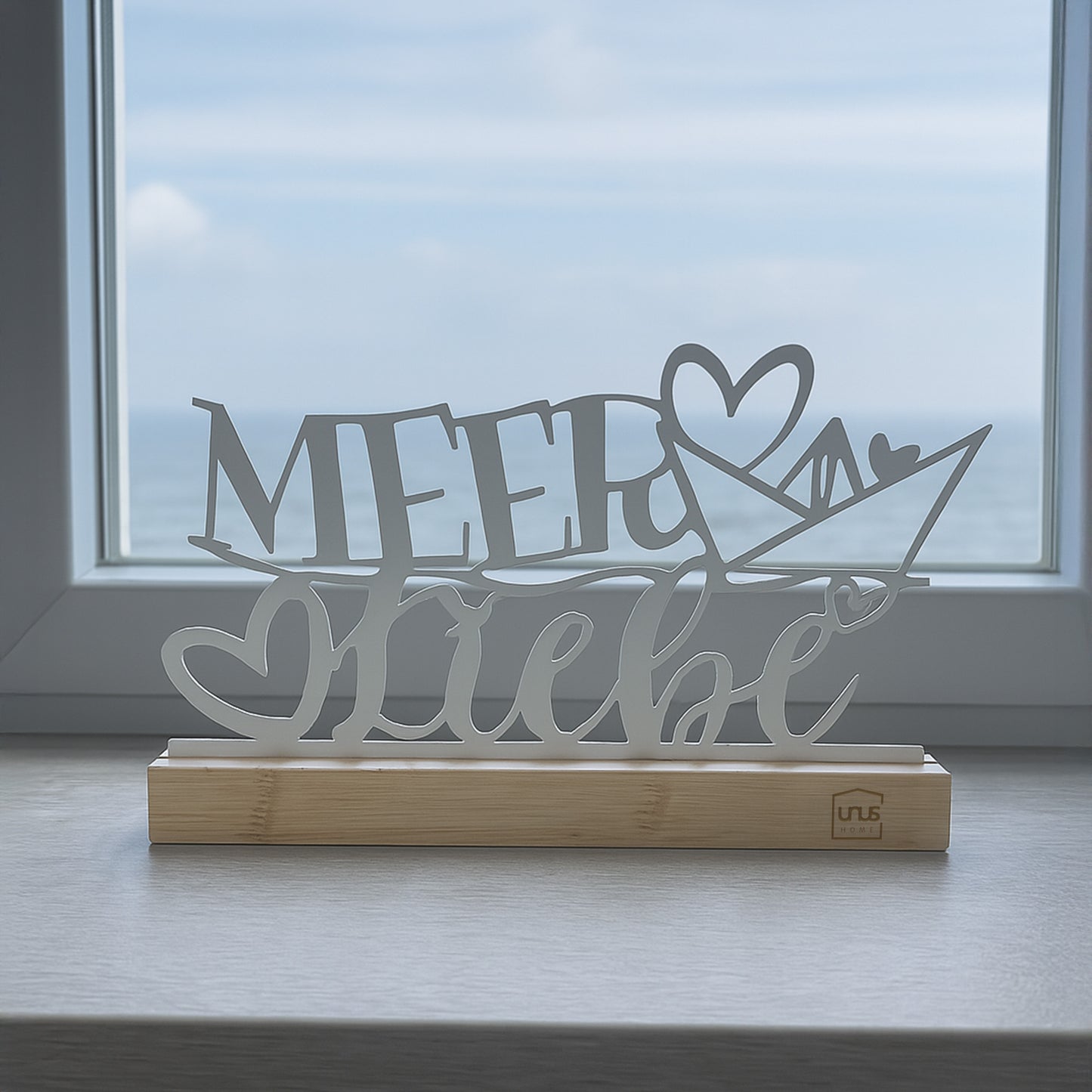 Weißer Aufsteller mit dem Schriftzug „Meer Liebe“ und Herz auf Holzsockel steht auf einer Fensterbank mit Blick auf das Meer und den Himmel.