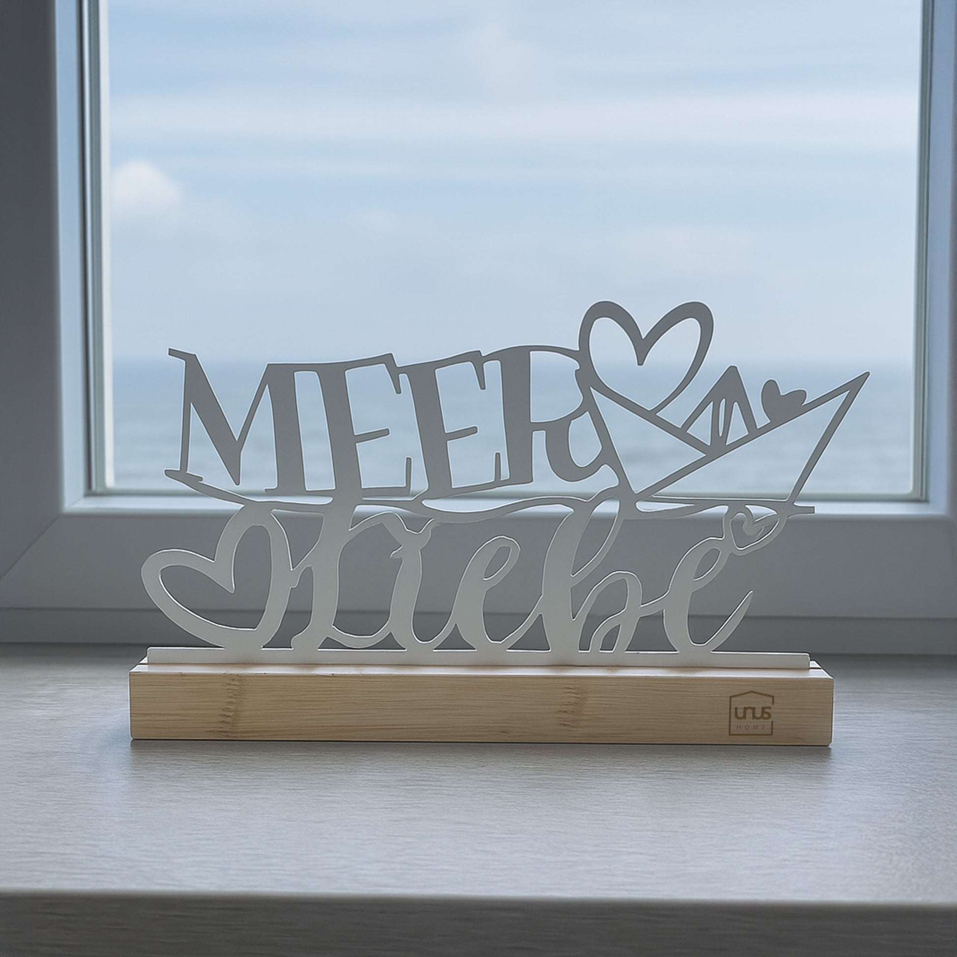 Weißer Aufsteller mit dem Schriftzug „Meer Liebe“ und Herz auf Holzsockel steht auf einer Fensterbank mit Blick auf das Meer und den Himmel.