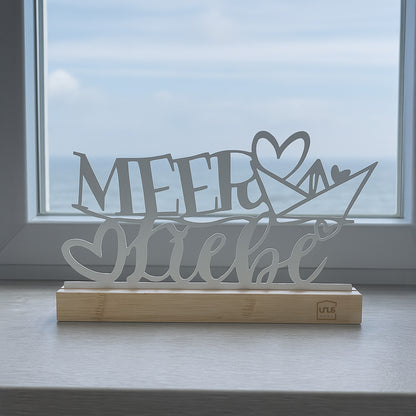 Weißer Aufsteller mit dem Schriftzug „Meer Liebe“ und Herz auf Holzsockel steht auf einer Fensterbank mit Blick auf das Meer und den Himmel.